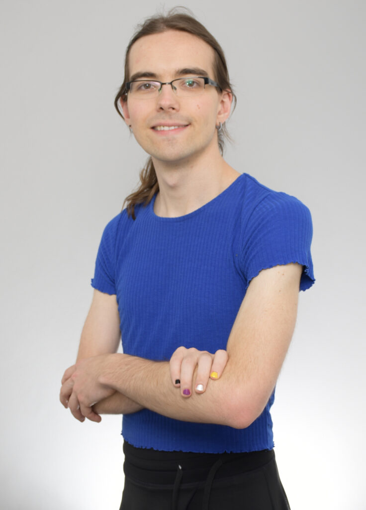 Fabi hat lange braune Haare, eine Brille und trägt ein blaues Oberteil mit gerafften Ärmeln. Die Nägel der linken Hand sind in den Farben der non-binary Flagge lackiert